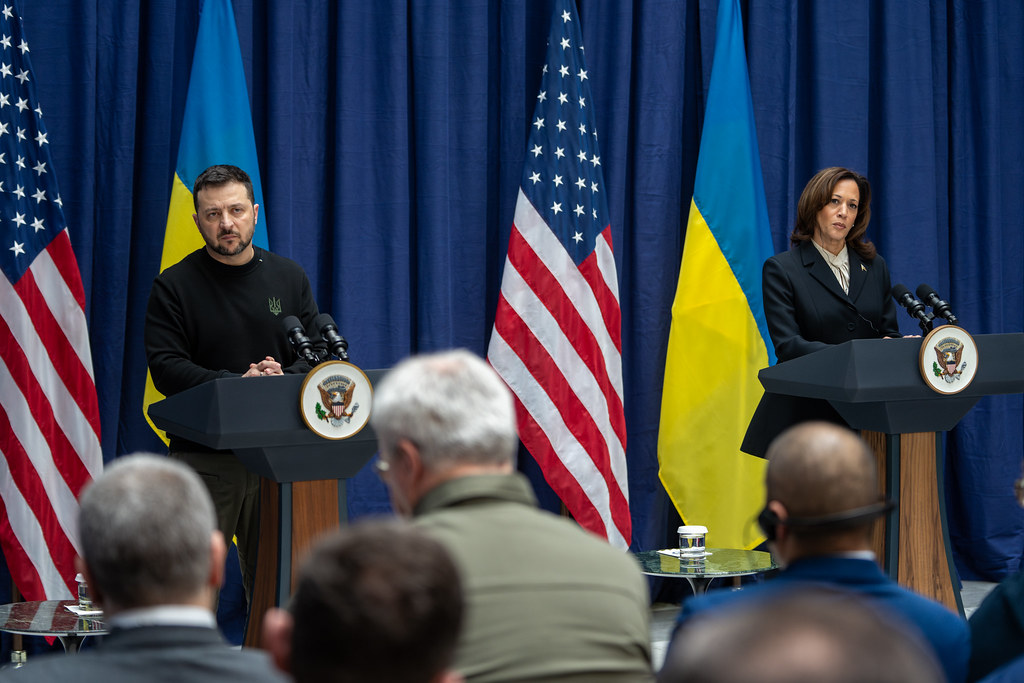 Vice President Kamala Harris and President Volodymyr Zelenskyy of Ukraine stand at separate podiums during a joint press conference at the Munich Security Conference. Behind them are the flags of the United States and Ukraine, and an audience listens attentively in the foreground.