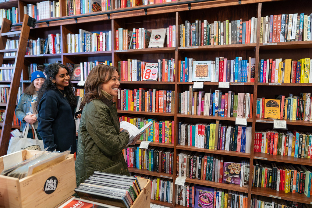 Vice President Kamala Harris smiles while browsing bookshelves at Bold Fork Books in Washington, DC. She is holding a book and is accompanied by two people in the cozy bookstore, which is filled with colorful books and a wooden ladder. The visit marks Small Business Saturday.