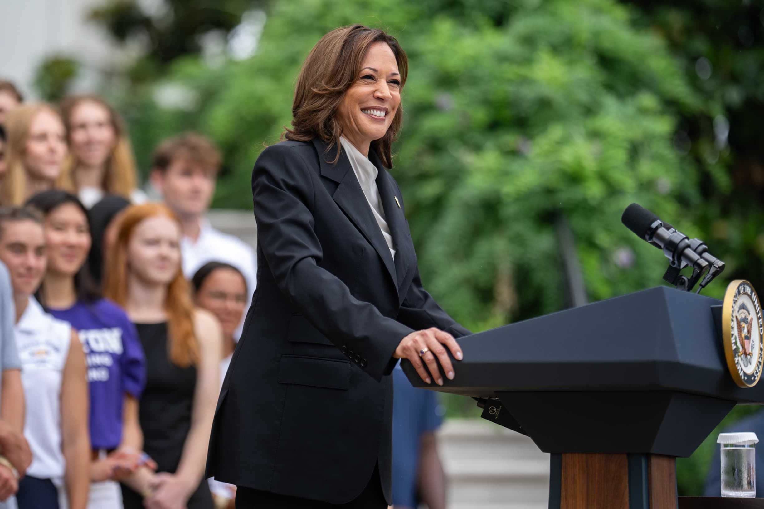 Vice President Kamala Harris smiles while speaking at a podium outdoors. She is surrounded by a group of young people standing in the background, with lush greenery framing the scene.