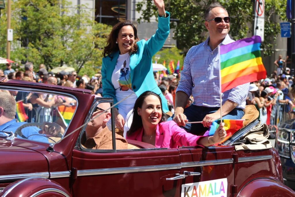 Vice President Kamala Harris rides in a convertible at a Pride Parade, smiling and waving to the crowd. She is joined by other participants, including her husband, Doug Emhoff, and her niece, Meena Harris, holding a rainbow Pride flag. The parade is vibrant, with a large crowd and rainbow decorations in the background.