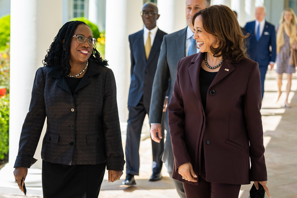 La vicepresidenta Kamala Harris y la jueza Ketanji Brown Jackson caminan juntas por la Columnata Oeste, sonriendo y conversando. Harris lleva un traje marrón y Jackson lleva un traje oscuro estampado. Otras personas las siguen en segundo plano mientras se dirigen a un evento que celebra la confirmación de Jackson para la Corte Suprema.