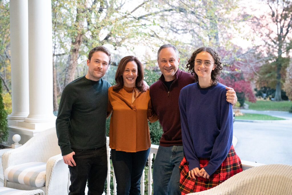 La vicepresidenta Kamala Harris, el segundo caballero Doug Emhoff y sus hijos Ella y Cole posan juntos en un porche decorado con muebles blancos. El grupo sonríe cálidamente, con árboles y un camino de entrada visibles al fondo.